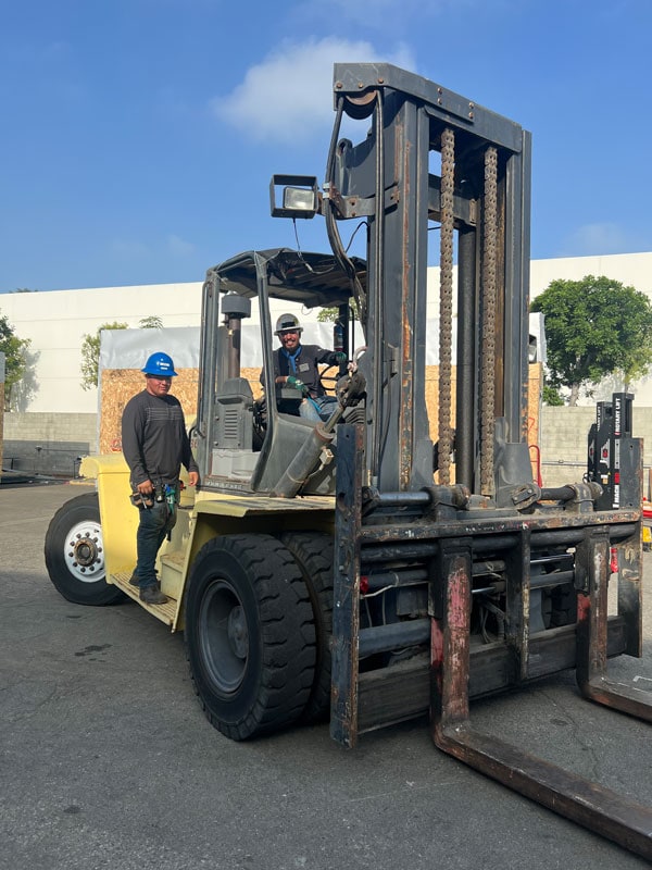 Two Crown employees smiling at the camera while riding on a forklift